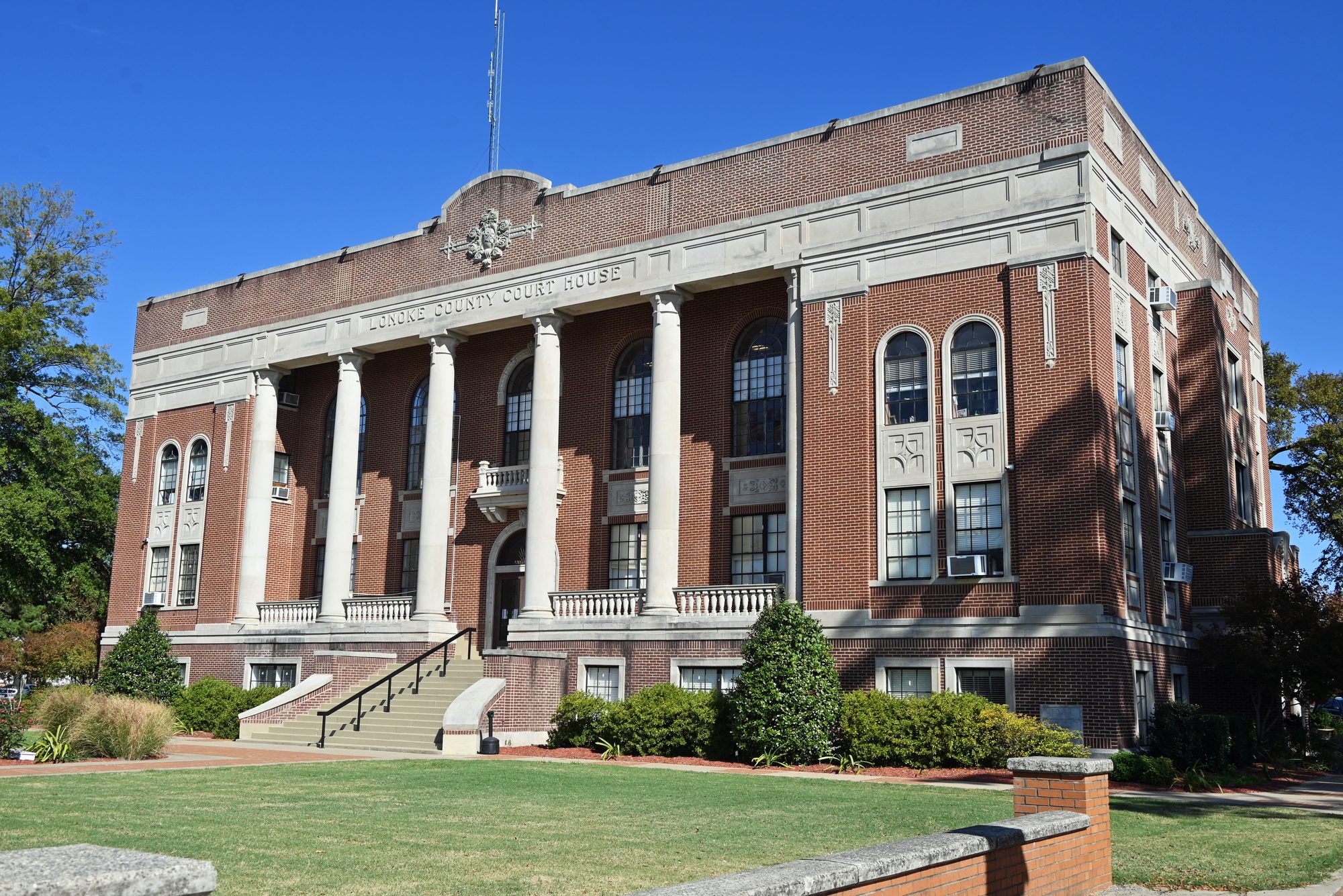 The Lonoke County Courthouse, a 1928 red-brick building with limestone columns, beneath a clear blue sky in Lonoke, Arkansas.