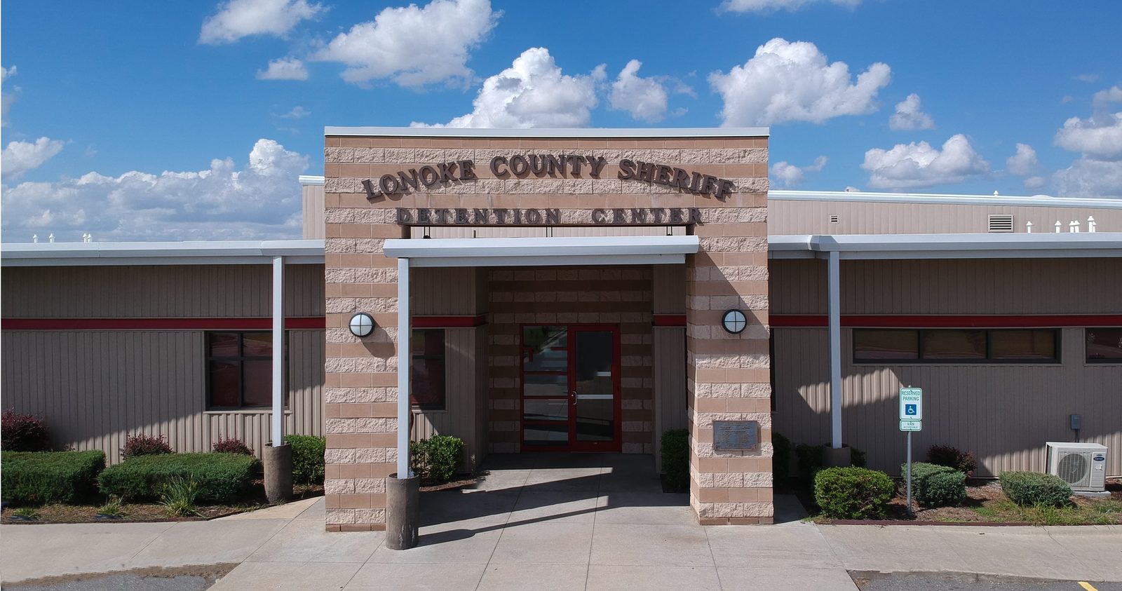 The front entrance of the Lonoke County Sheriff Detention Center, a tan block building beneath a blue sky.